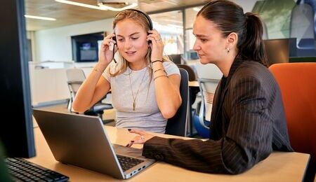  Klantcontact collega's Twee collega's zitten naast elkaar aan het bureau. Een collega zit achter haar laptop en heeft een headset op. De andere collega lijkt mee te kijken/haar in te werken.