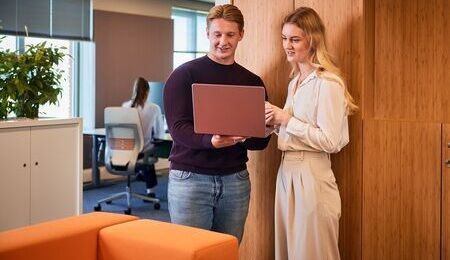 Twee collega's staan voor een hoge kast op kantoor en kijken allebei naar een laptopscherm die een collega vasthoudt.