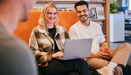  VvAA Collega's Twee collega's zitten op een oranje bank. De vrouw heeft een laptop op haar schoot en kijkt lachend de camera in. De man kijkt naar het beeldscherm en lacht.
