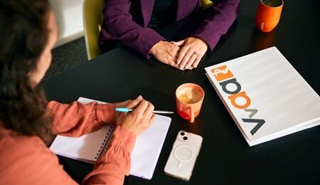 Een zwarte tafel waar je twee mensen aan ziet zitten. Op de tafel liggen documenten, een pen, een telefoon en een oranje koffiemok.